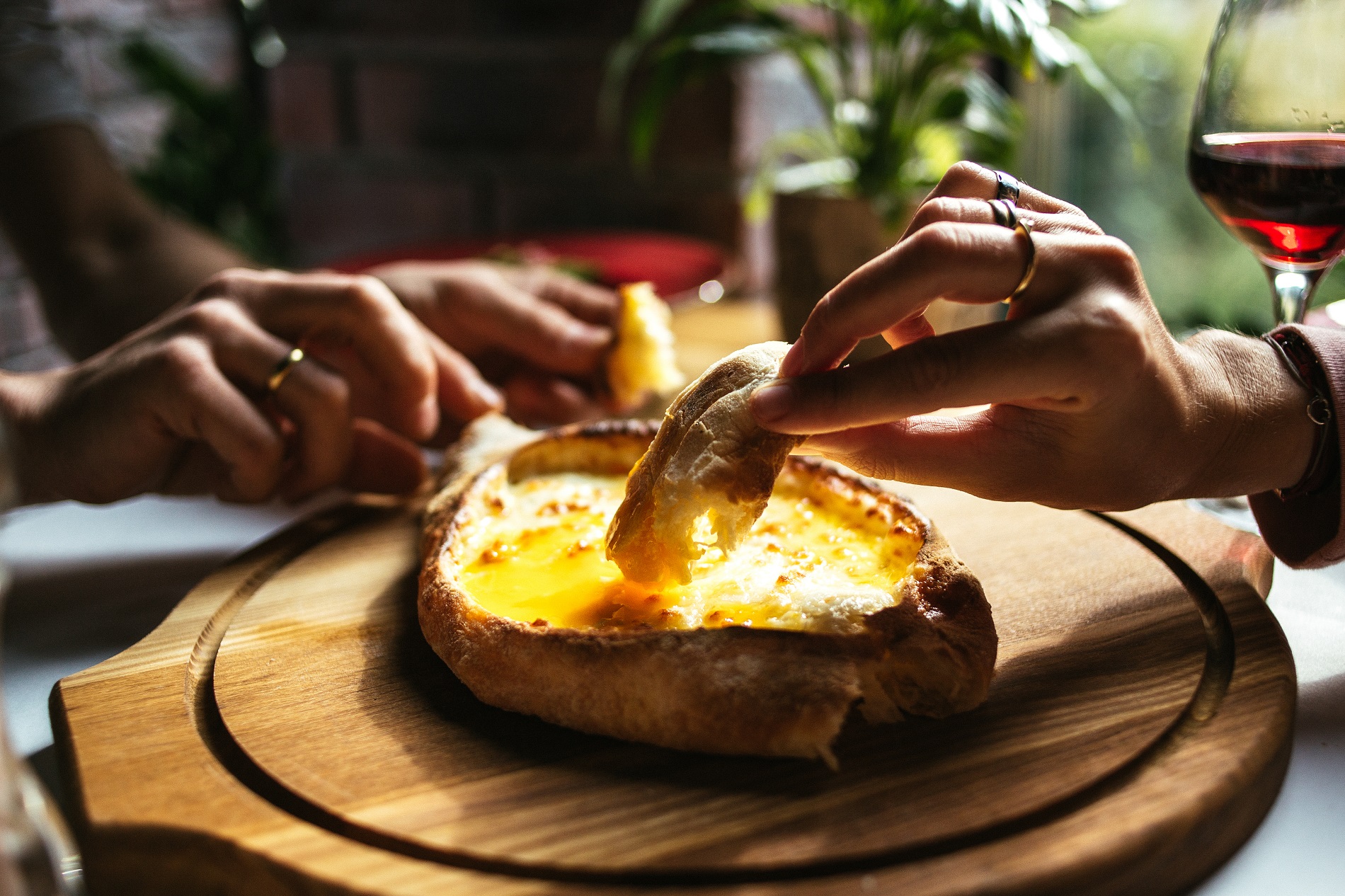 This captivating photo showcases a beautifully set table adorned with a delectable spread of authentic Georgian cuisine. A family gathers around, savoring the iconic Khachapuri Adjarian Style, a cheese-filled bread masterpiece. The Khachapuri, placed on a rustic wooden tray, takes center stage, enticingly inviting the viewers to indulge. A woman's hand, adorned with elegant rings, delicately holds a mouthwatering piece of Khachapuri, capturing the tactile experience. In the background, the presence of a man's hands and a glass of red wine enhances the atmosphere of conviviality and enjoyment. This photo encapsulates the essence of a traditional Georgian family meal, where the warm flavors of Khachapuri and the richness of red wine create a memorable dining experience.